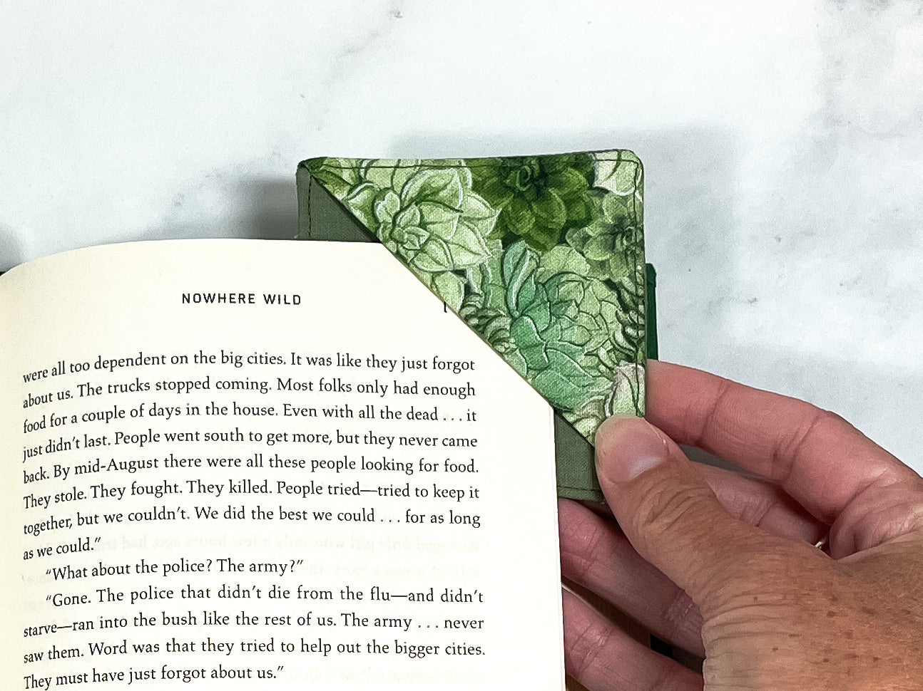 A handmade corner bookmark made from a fun succulent plant themed cotton fabric in shades of green.  The page marker is square and is being shown sliding over the corner of a page in a book.