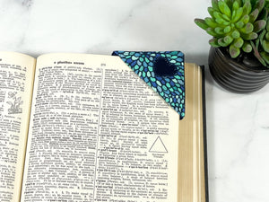 A handmade corner bookmark made from a fun mosaic and heart themed cotton fabric in shades of aqua and navy blue.  The page marker is square and is being shown on the corner of a book with a succulent plant off to the side.
