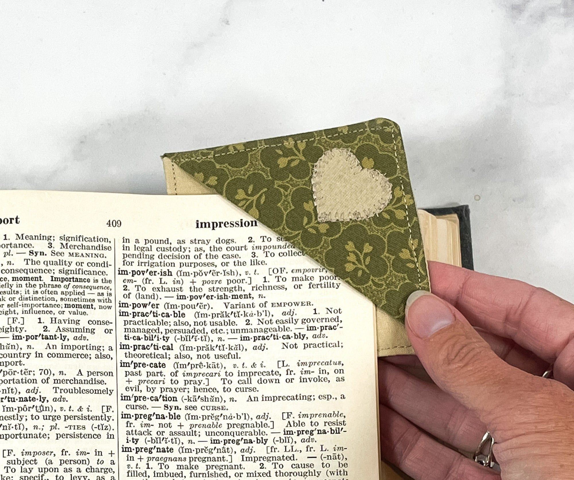 A handmade corner bookmark made from a fun rustic country floral and heart themed cotton fabric in shades of olive green and tan.  The page marker is square and is being shown sliding over the corner of a page in a book.