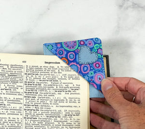 A handmade corner bookmark made from a fun modern blue themed cotton fabric in shades of blue.  The page marker is square and is being shown sliding over the corner of a page in a book.