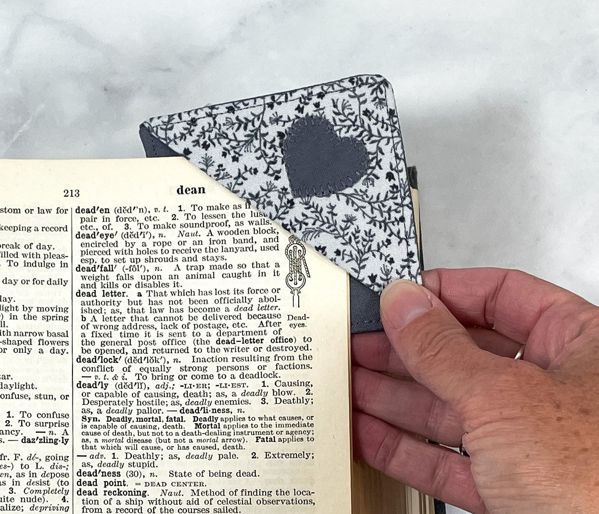 A handmade corner bookmark made from a fun flower and heart themed cotton fabric in shades of gray.  The page marker is square and is being shown sliding over the corner of a page in a book.