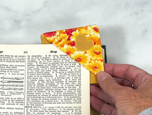 A handmade corner bookmark made from a fun floral and heart themed cotton fabric in shades of yellow, orange and red.  The page marker is square and is being shown sliding over the corner of a page in a book.