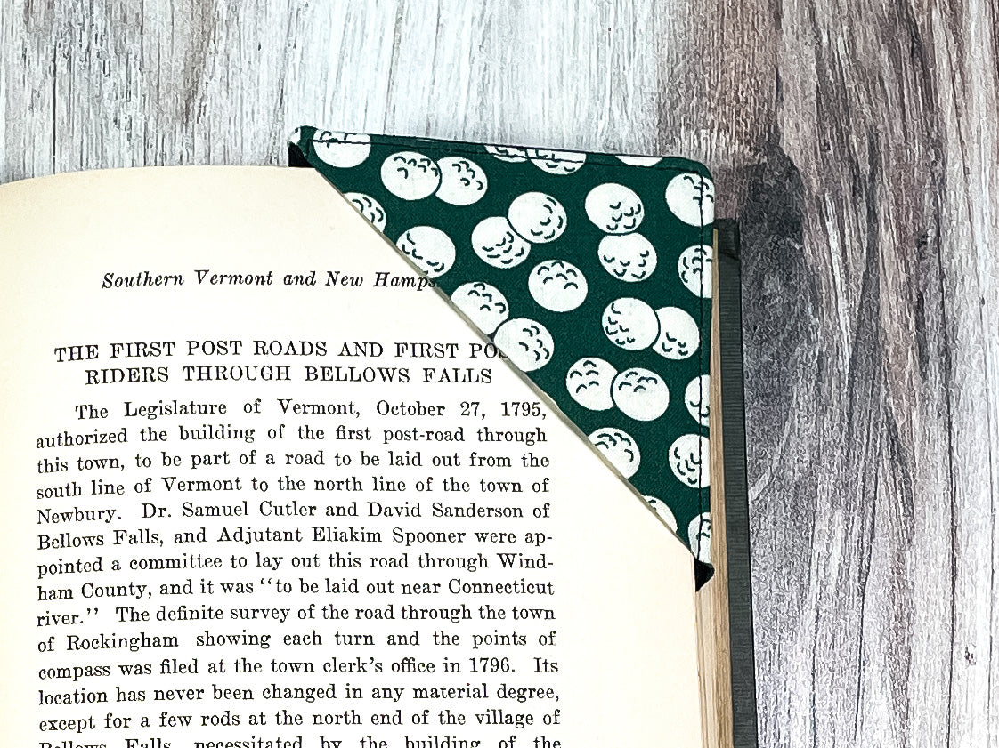 A handmade corner bookmark made from a fun golf themed cotton fabric in shades of black, green and white.  The page marker is square and is being shown sliding over the corner of a page in a book.