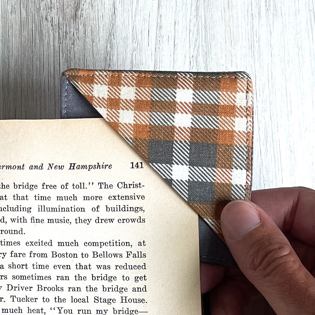 A handmade corner bookmark made from a fun checkered plaid themed cotton fabric in shades of gray, brown and white.  The page marker is square and is being shown sliding over the corner of a page in a book.