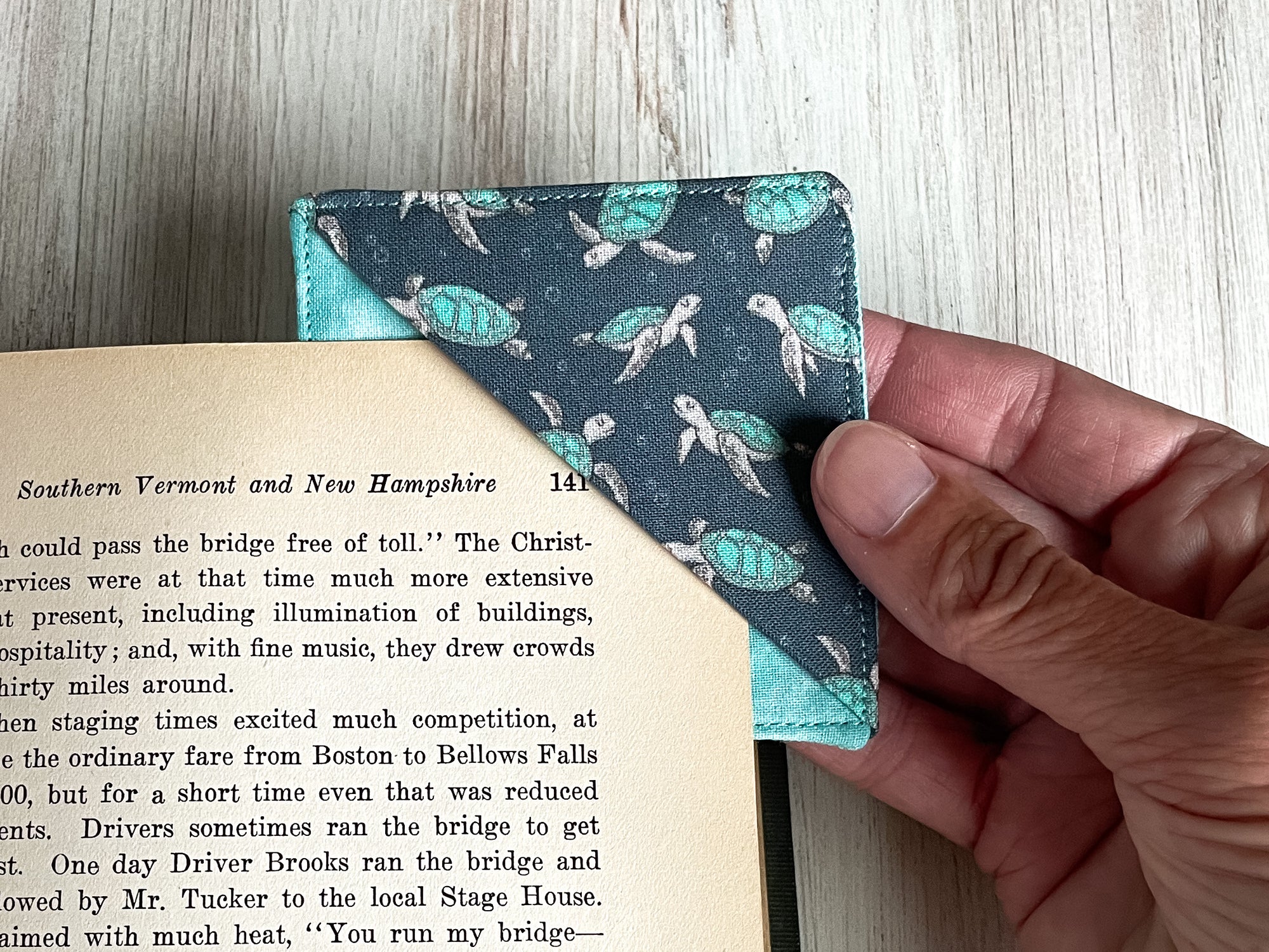 A handmade corner bookmark made from a fun turtle themed cotton fabric in shades of aqua and navy blue.  The page marker is square and is being shown sliding over the corner of a page in a book.