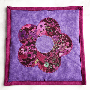 A single floral themed potholder made with a flower applique pattern.  The fabrics used are purple and mauve and will add a nice cottagecore feel to your kitchen island or dining room table.
