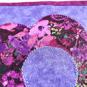 A single floral themed potholder made with a flower applique pattern.  The fabrics used are purple and mauve and will add a nice cottagecore feel to your kitchen island or dining room table.